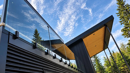 Matte black side-mount fascia spigots securing frameless glass balcony panels on a modern home with dark exterior cladding.